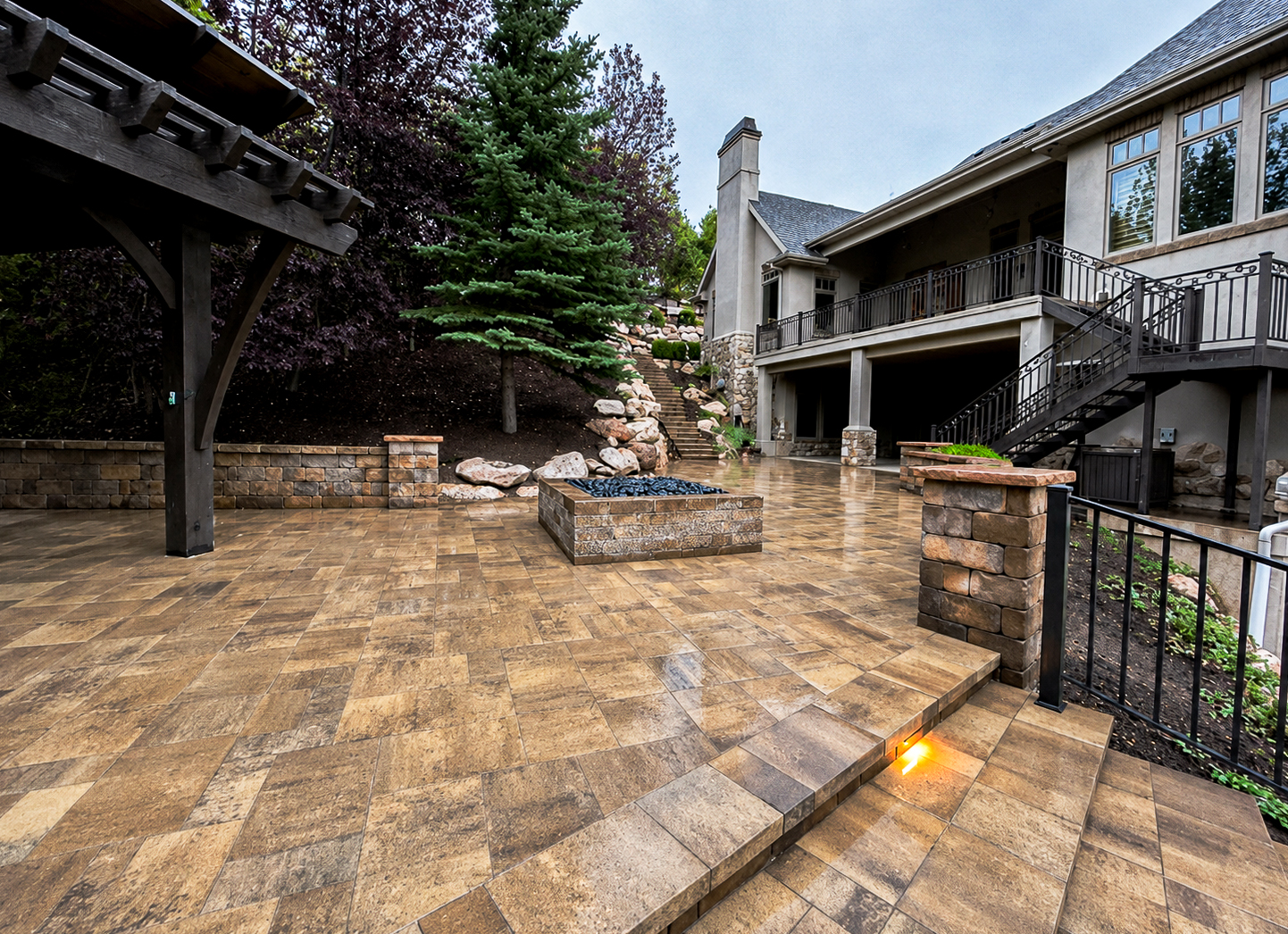 Retaining Walls in Utah Multi-level paver patio at dusk with a square fire pit, stone pillars and a wood pergola, retaining walls by Empire Pavers in Utah
